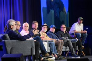 From left to right, during D23’s “A Beautiful Tomorrow... 60 Years of the World’s Fair” panel, are Leonard Maltin, Pete Docter, Tom Fitzgerald, Ted Ryan, Ramsey Avery, Kevin Feige, Jon Favreau, and Michael Giacchino. They’re sitting on gray sofa chairs, and the background features a blue curtain.