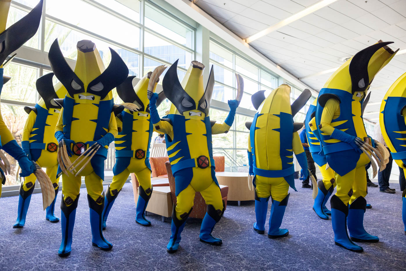 A group of cosplayers dressed as “Peelverines,” a creative Fortnite-related mashup of Peely the banana and Wolverine, at D23: The Ultimate Disney Fan Event 2024. They’re striking dynamic poses on a purple carpeted floor, with large windows in the background revealing the interior space where the photo is taken.