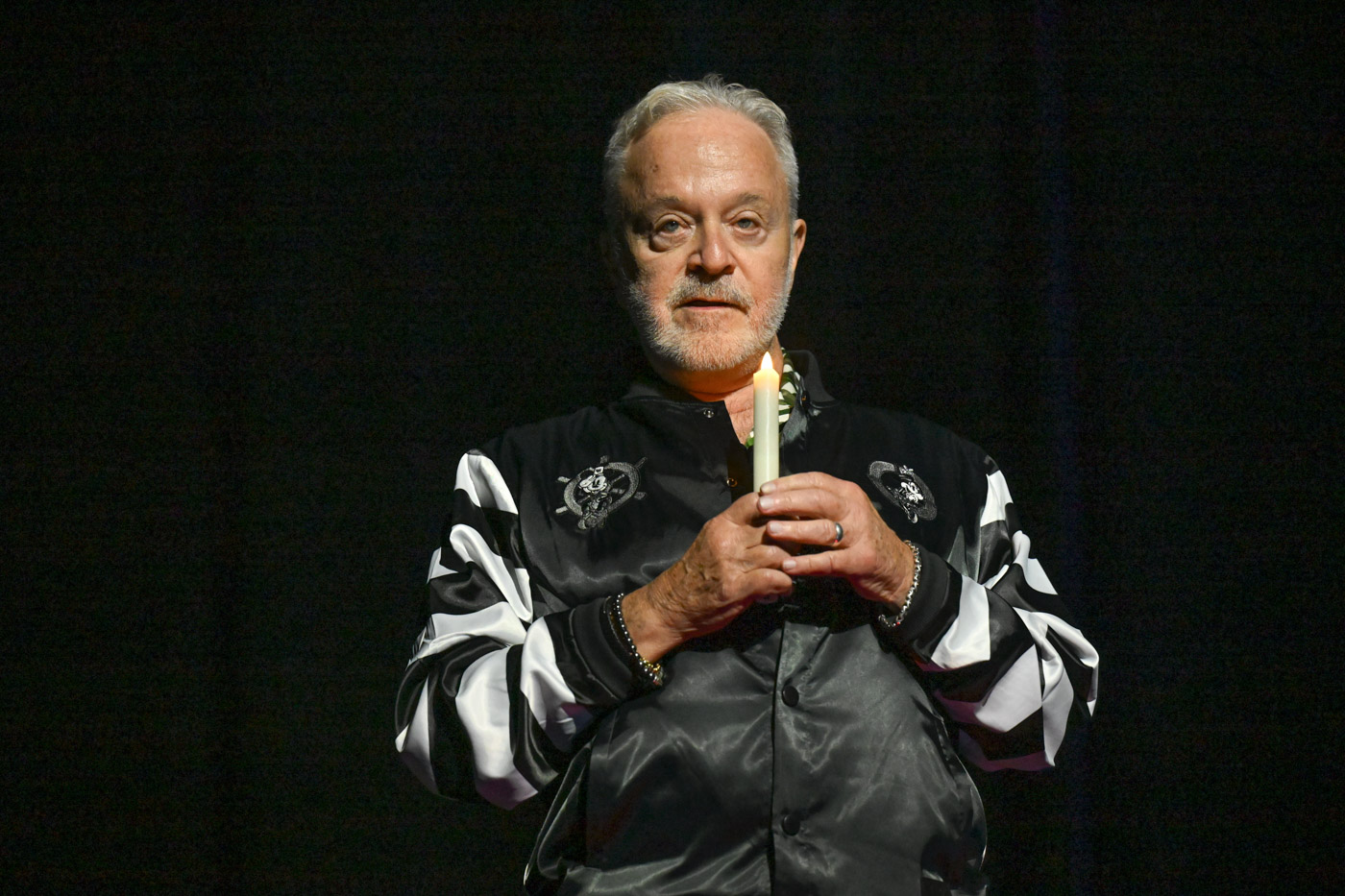 Jim Cummings is standing in front of a black background, holding a lit candle with his hands. He is wearing a black and white Disney bomber jacket. 