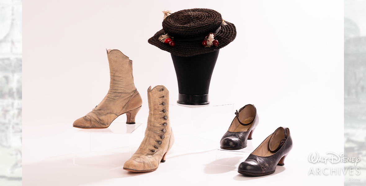 Several pieces of Mary Poppins’ wardrobe, all worn by Disney Legend Julie Andrews are shown. From left, the items include Mary’s since-painted-white “Jolly Holiday” boots with silver metal buttons, her Edwardian-style black straw hat with plastic red cherries and white silk daisies, and her blue leather shoes on the right.