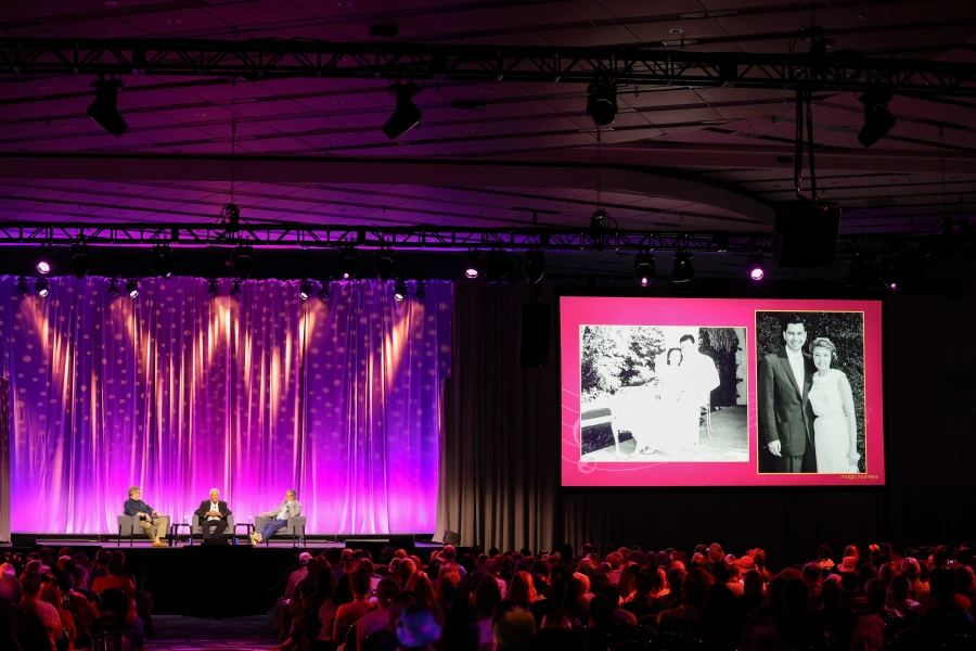 From left to right, host Tim O’Day, Jeff Sherman, and Greg Sherman are sitting in gray sofa chairs on a stage during the “Magic Journeys: Memories of the Sherman Brothers” panel at D23: The Ultimate Disney Fan Event. Fans fill the audience. The background features a blue stage curtain, with purple, pink, and yellow lights. To the panel’s right is a screen projecting black and white photos of the Sherman Brothers’ wedding days. 