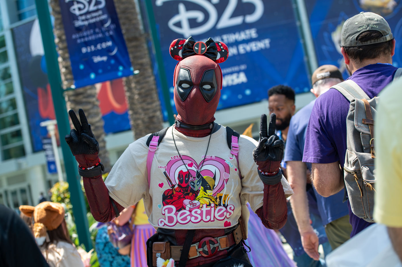 A fan dressed as Deadpool is posing outside the Anaheim Convention Center during D23: The Ultimate Disney Fan Event. They’re wearing a Deadpool costume with a crop graphic tee featuring Deadpool and Wolverine with “Besties” written on it, along with Minnie Mouse ears. The fan is making peace signs with their hands.