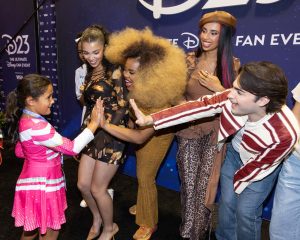 At a photo opportunity (from left to right), Malia Baker, Dara Reneé, Kylie Cantrall, and Joshua Colle from Descendants: The Rise of Red and ZOMBIES 4 are interacting with a young fan at D23: The Ultimate Disney Fan Event. Reneé and Colle are high-fiving the young fan.