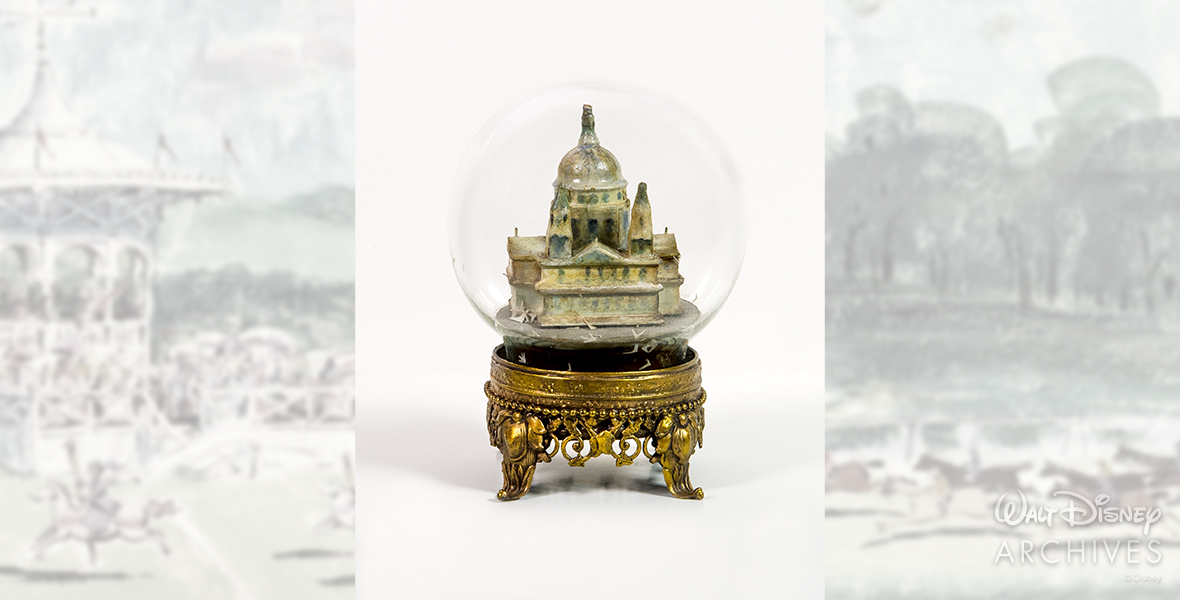 Shown is a snow globe prop featuring St. Paul’s Cathedral. The globe has been drained of water and features a painted cathedral within a circular dome. There are paper birds at the bottom of the globe, which has an intricate golden metal base.