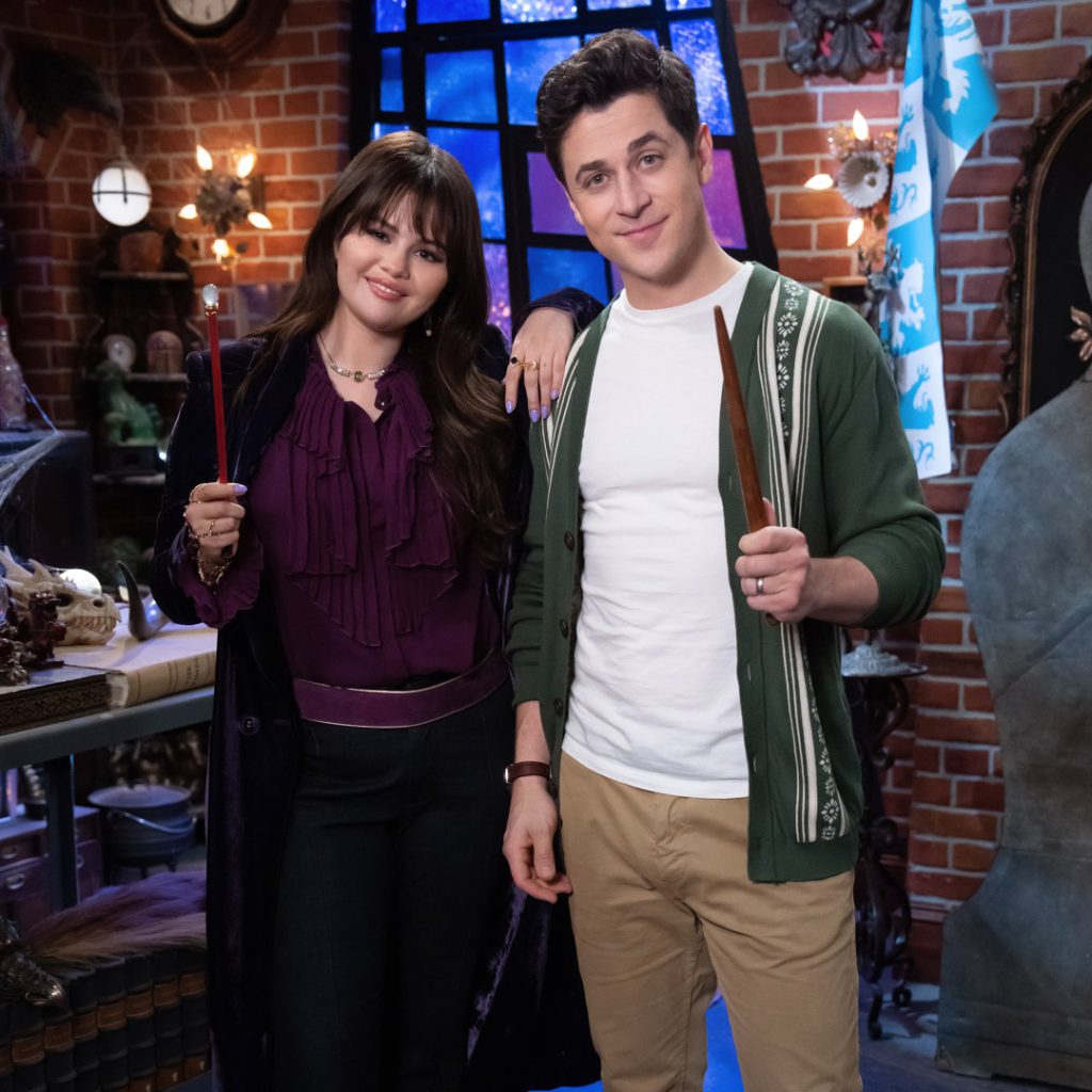 Selena Gomez and David Henrie stand side by side, holding wands in a magical, wizard-themed room. The setting includes brick walls, mystical objects, and a window with a glowing blue and purple light. Selena is dressed in a dark purple blouse and black pants, while David is wearing a green cardigan over a white shirt and beige pants.