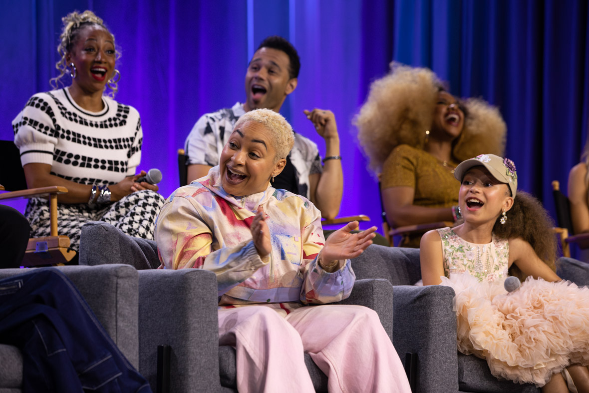 Raven-Symoné (center), is seen on stage with Mykal-Michelle Harris (to her right) and (l-r behind her) Monique Coleman, Corbin Bleu, and Dara Reneé, during the “Stay Tuned: You’re Watching Disney Channel” panel at D23: The Ultimate Disney Fan Event. They’re all laughing and smiling at someone off camera to the left.