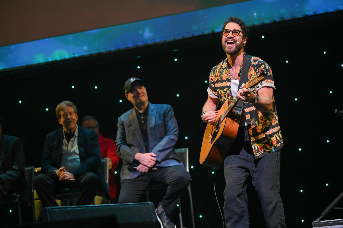 Darrin Criss on stage at “Jolly Holiday: A D23 Musical Celebration of Richard M. Sherman” during D23: The Ultimate Disney Fan Event 2024. He’s holding a guitar and singing. To his left are Marvel Studios president Kevin Feige and Disney Legend Tony Baxter.