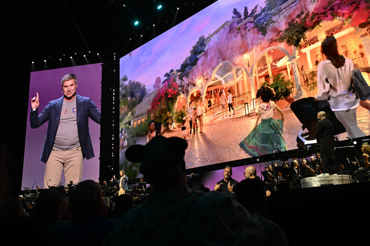 On Saturday, August 10, 2024, at the Honda Center, BRUCE VAUGHN took the stage during the DISNEY EXPERIENCES marquee showcase. A large concept drawing showcasing an Encanto-themed addition to Disney’s Animal Kingdom is displayed behind him.