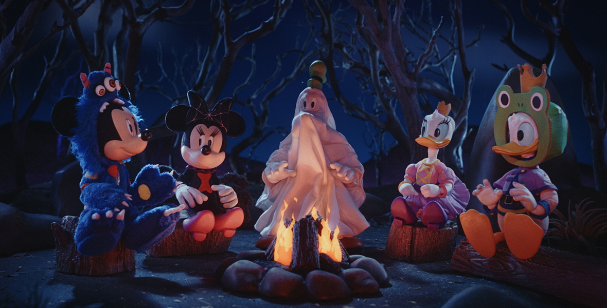 Sitting around a campfire in their Halloween costumes are the stop-motion puppets for Mickey Mouse, Minnie Mouse, Goofy (his face and body obscured by his white sheet of a ghost costume), Daisy Duck, and Donald Duck. In this scene from Mickey’s Spooky Stories, it’s nighttime and creepy trees are behind the line of characters, who are all sitting on boulders. The fire burns within a circle of smaller stones.