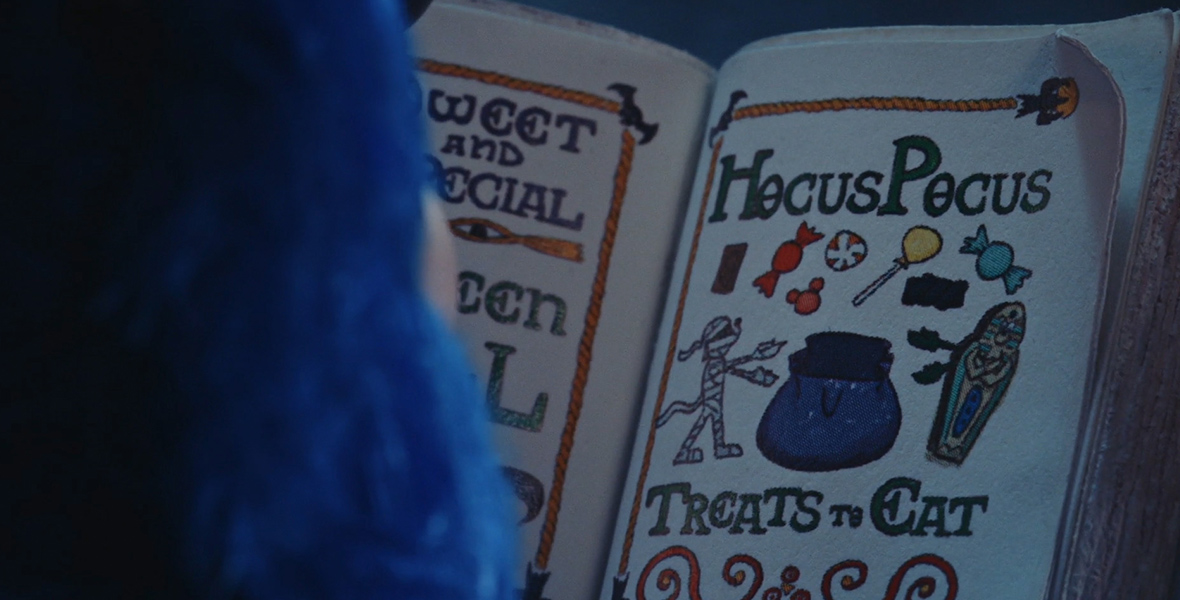 In this scene from Mickey’s Spooky Stories, a close-up view of an open spell book shows the words Hocus Pocus at the top of a page. At the bottom of the page are the words Treats to Eat. In between are cartoon-like drawings of treats, a mummy, a trick-or-treat sack, and more. One of the little treat icons is shaped like Mickey’s head.