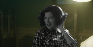 In a scene from Agatha All Along, Agatha Harkness (Kathryn Hahn) wears a retro, short hairstyle and a checkered shirt. She is smiling mischievously and posing with one hand in her hair.