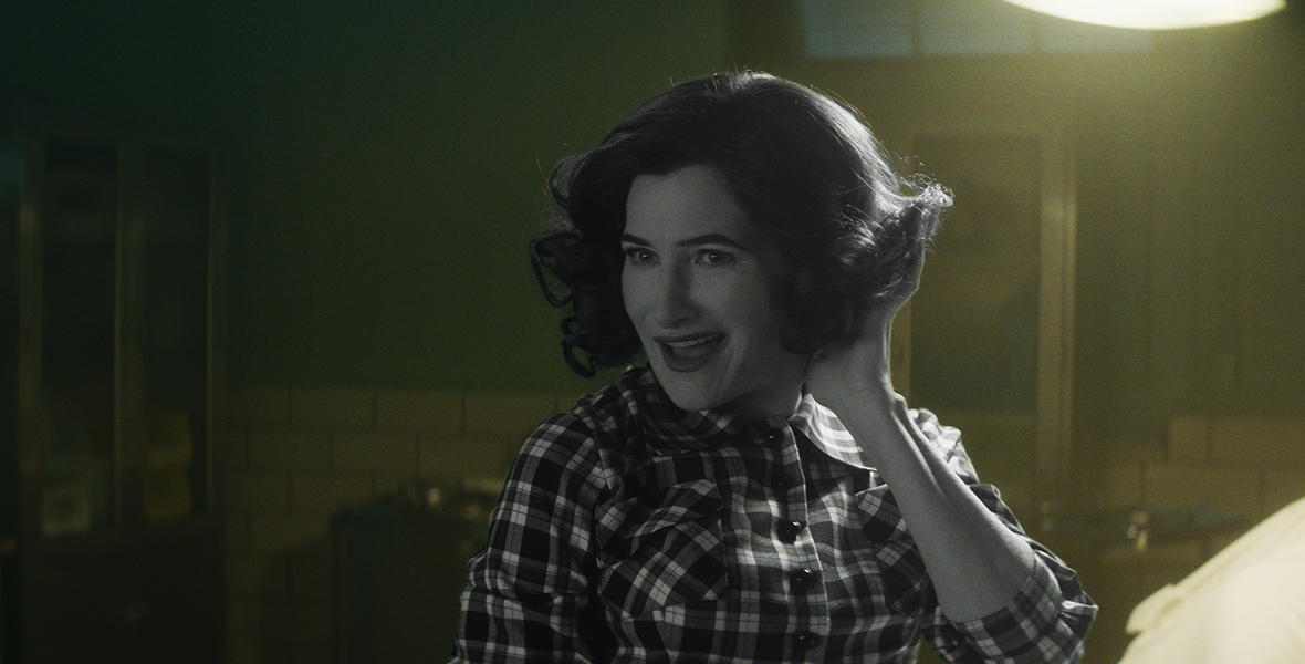 In a scene from Agatha All Along, Agatha Harkness (Kathryn Hahn) wears a retro, short hairstyle and a checkered shirt. She is smiling mischievously and posing with one hand in her hair. 