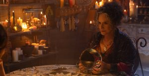 In a scene from Agatha All Along, Lilia Calderu (Patti LuPone) sits on a table, gazing at Agatha Harkness (Kathryn Hahn) while holding a crystal ball. The background features lit and unlit candles, as well as paintings. The table is draped with a white and black patterned tablecloth. Lilia wears a black and purple/pink patterned robe.