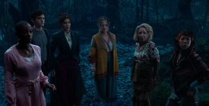 In a scene from Agatha All Along, (from left to right), Jennifer Kale (Sasheer Zamata), Teen (Joe Locke), Agatha Harkness (Kathryn Hahn), Lilia Calderu (Patti LuPone), Mrs. Hart/ Sharon Davis (Debra Jo Rupp), and Alice Wu-Gulliver (Ali Ahn) stand in a dark forest surrounded by dead leaves. They are wearing a mix of modern and eclectic clothing, and their expressions are tense, with everyone seemingly prepared for something ominous.