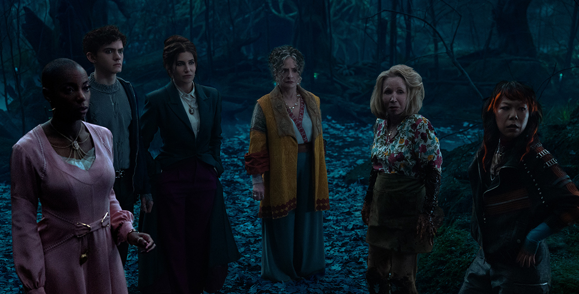 In a scene from Agatha All Along, (from left to right), Jennifer Kale (Sasheer Zamata), Teen (Joe Locke), Agatha Harkness (Kathryn Hahn), Lilia Calderu (Patti LuPone), Mrs. Hart/ Sharon Davis (Debra Jo Rupp), and Alice Wu-Gulliver (Ali Ahn) stand in a dark forest surrounded by dead leaves. They are wearing a mix of modern and eclectic clothing, and their expressions are tense, with everyone seemingly prepared for something ominous. 