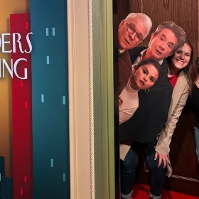 Four fans enjoy the Only Murders in the Building photo op featuring Charles, Oliver, and Mabel peeking out of a doorway.