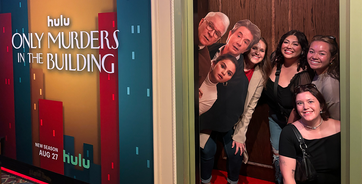 Four fans enjoy the Only Murders in the Building photo op featuring Charles, Oliver, and Mabel peeking out of a doorway.