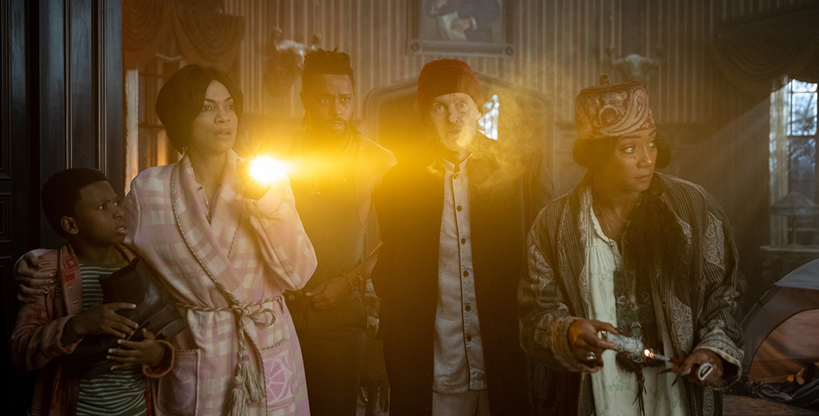 In a scene from Haunted Mansion (2023), from left to right, Travis (Chase Dillon), Gabbie (Rosario Dawson), Ben Matthias (LaKeith Stanfield), Father Kent (Owen Wilson), and Harriet (Tiffany Haddish) stand in sleeping attire—pajamas and robes—with confused expressions as they look to their left. Gabbie holds a flashlight in her left and Travis with her right. The background shows a room with a tent on the floor, two windows, and a painting in the middle of the far wall.