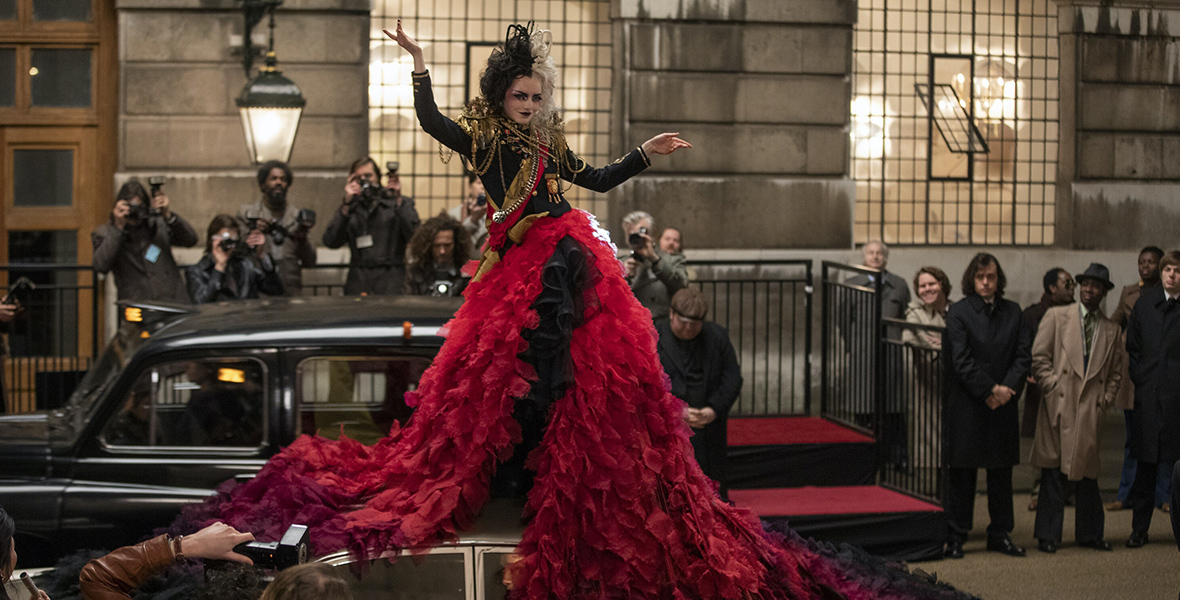 In a scene from Cruella, Emma Stone as Cruella sits on top of a car, posing for paparazzi. She wears a maxi dress with a red and black tulle ball gown bottom and a black and gold long-sleeve top with gold shoulder pads. Her hair is half black, half white. In the background, another car and more paparazzi stand outside a gray building.