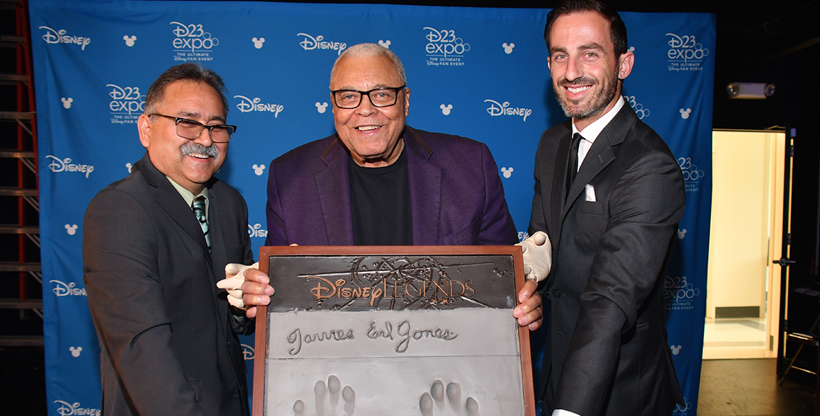 James Earl Jones (center) holds up his cement handprints and autograph after being inducted as Disney Legend in 2019. He is flanked by a man on each side.