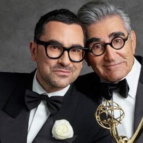 Alt Text: Dan Levy (left) rests his head on Eugene Levy (right), who is holding an Emmy.