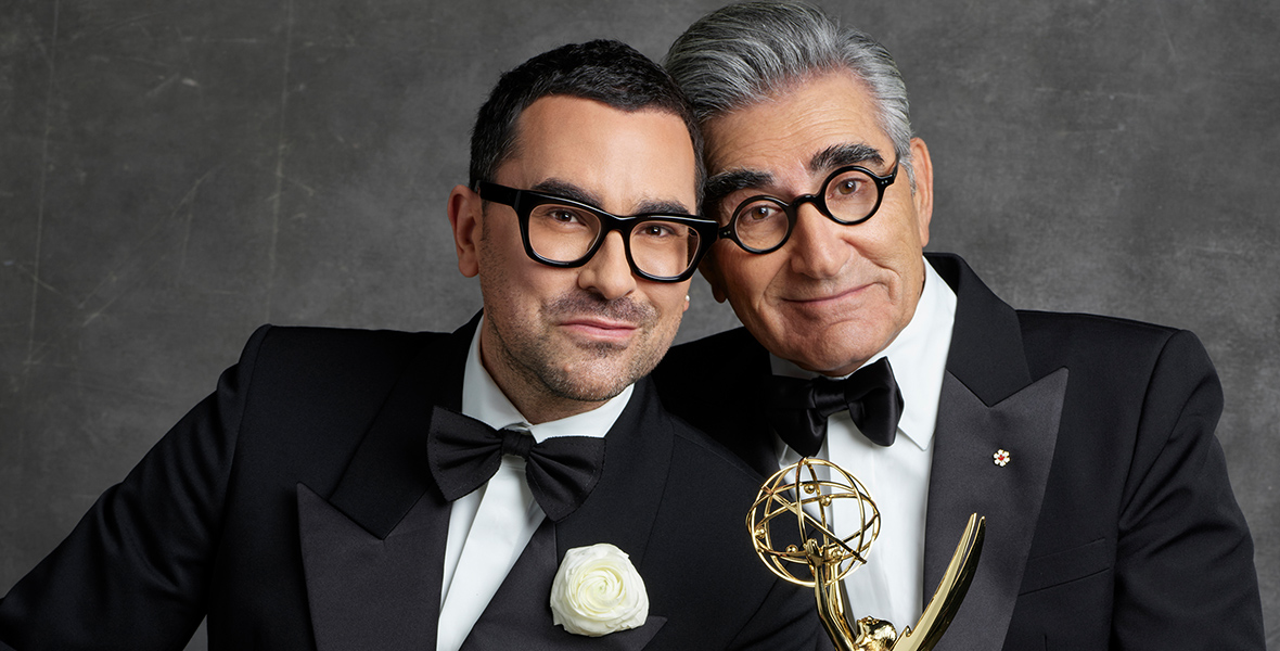 Alt Text: Dan Levy (left) rests his head on Eugene Levy (right), who is holding an Emmy.