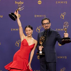 A celebratory image featuring two actors holding Emmy Awards at the Emmy Awards ceremony. On the left is Anna Sawai in a striking red gown joyfully raises her Emmy high. On the right is Hiroyuki Sanada in a classic dark suit proudly holds his Emmy.