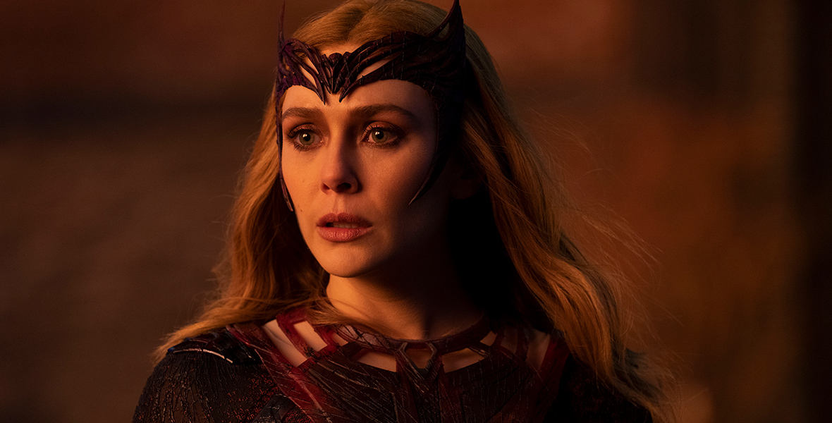 In a scene from Doctor Strange, Wanda Maximoff (Elizabeth Olsen), also known as the Scarlet Witch, dons a red long-sleeve suit with square cutouts at the neckline. She wears a horned headpiece that frames her face. The background is a blur of warm brown and orange tones.