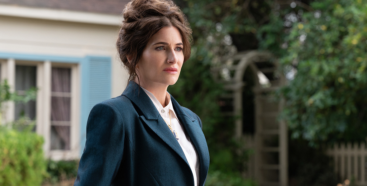 In a scene from Agatha All Along, Agatha Harkness (Kathryn Hahn) wears a navy blue fitted suit over a collared white shirt, and a gold pendant necklace. Her hair is in an updo, and the background features the exterior of a house and its garden.