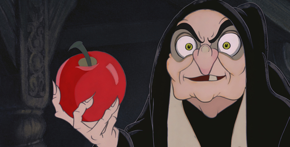 In a scene from Snow White and the Seven Dwarfs (1937), the Evil Queen (Lucille La Verne) is disguised as an old witch. She wears a black cloak and extends her right hand, which holds a crimson red apple. The background depicts a dark, wooden interior.