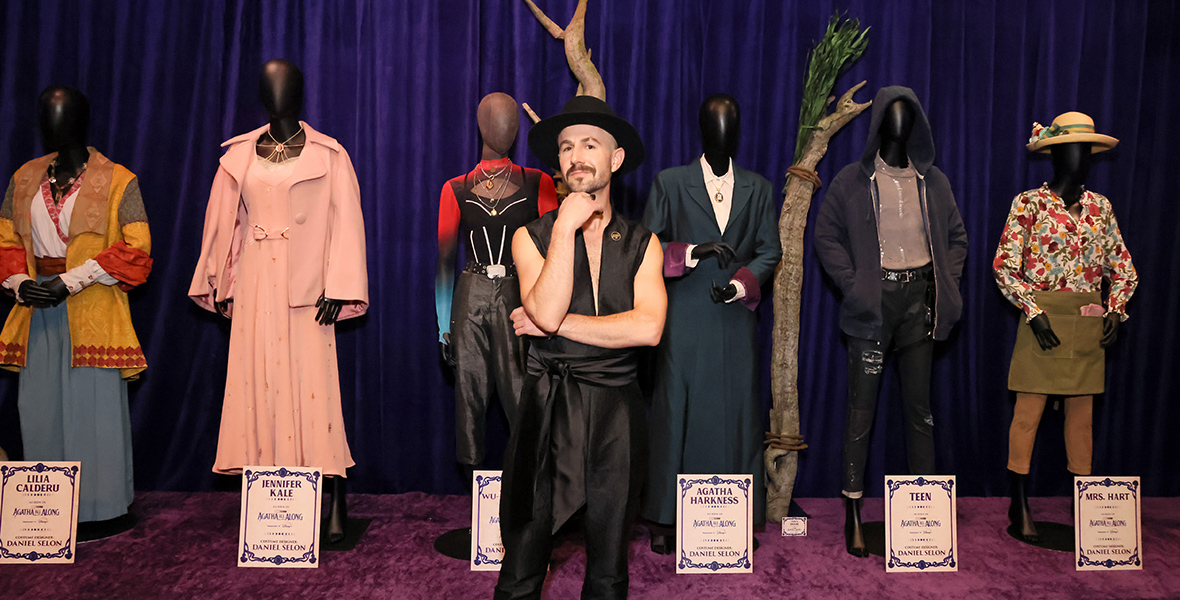 In a photo from the Agatha All Along premiere, costume designer Daniel Selon stands before six costumes displayed on mannequins, each labeled with the character's name. Selon is dressed in a black vest and matching pants, complemented by a black top hat. The background features a dark purple curtain and two thick branches, adding a touch of drama to the scene.