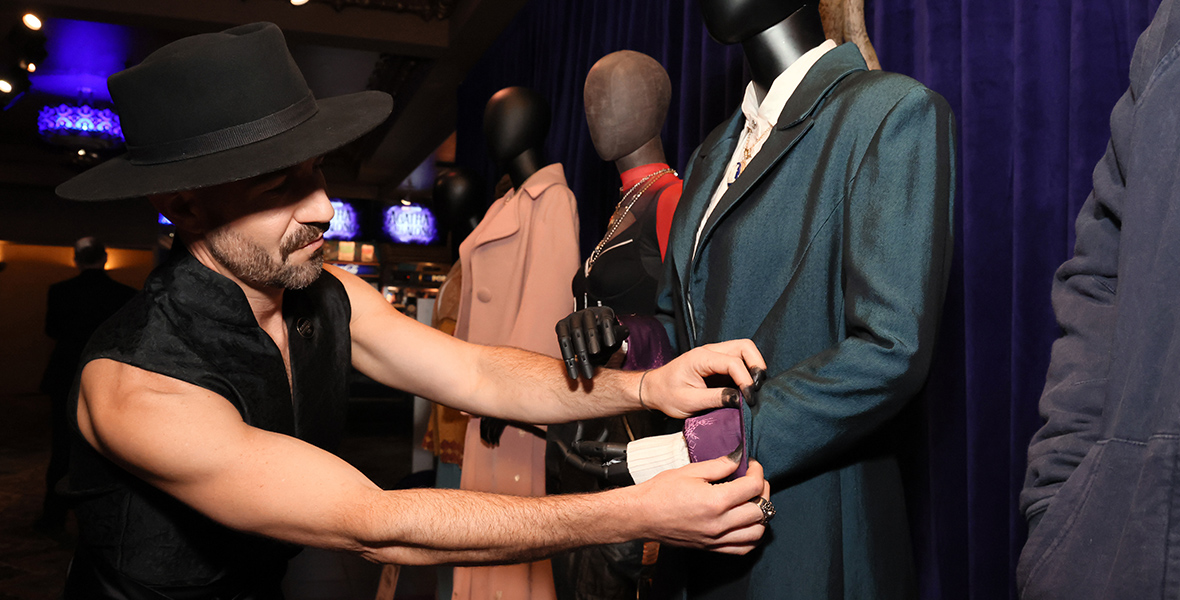 In a photo from the Agatha All Along premiere, costume designer Daniel Selon stands in front of a mannequin dressed in a navy blue, cinched long-sleeve coat over a white shirt. He is adjusting the coat’s left cuff to reveal a vibrant purple lining. Selon is outfitted in a black vest and a black top hat. The background showcases additional mannequins in costumes and is set against a dark purple curtain.