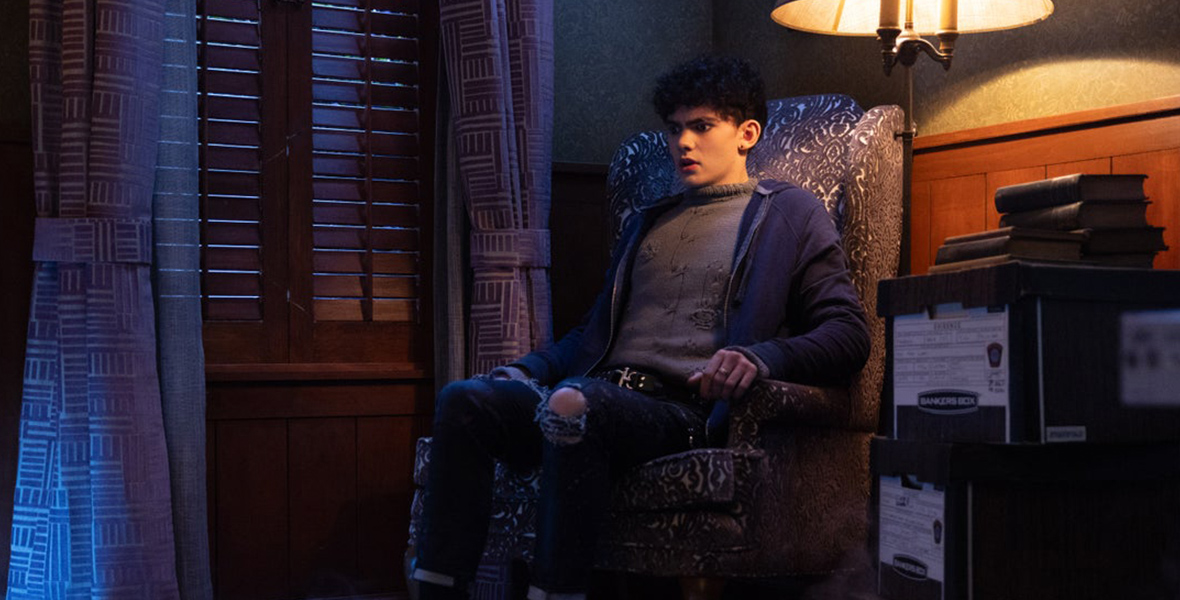 In a scene from Agatha All Along, Teen (Joe Locke) sits on a wingback chair with black and white patterns, his face registering a shocked expression. He's dressed in a gray sweater layered under a navy-black zip-up cardigan, paired with black ripped jeans and a black belt with a silver buckle. A mist creeps across the room’s floor, while the rest of the space features a warm-lit lamp, books, and curtains.