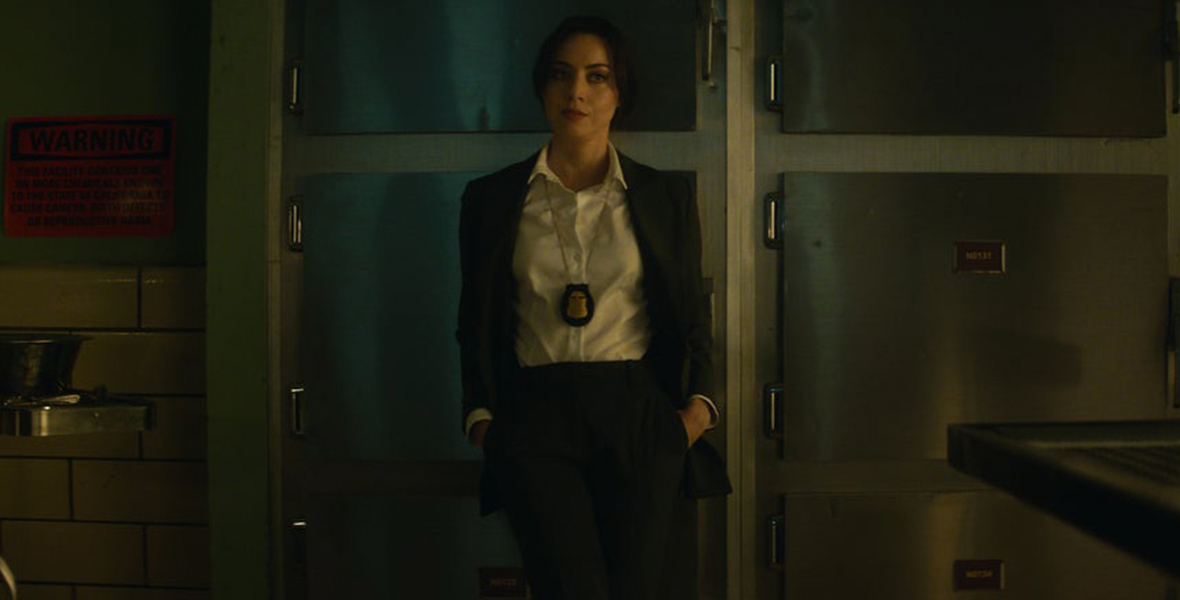 Rio Vidal in a scene from Agatha All Along, Rio Vidal (Aubrey Plaza) is dressed in a dark gray suit and matching pants over a white shirt, with a police badge hanging around her neck. She stands in a morgue, with metallic cabinets visible in the background.