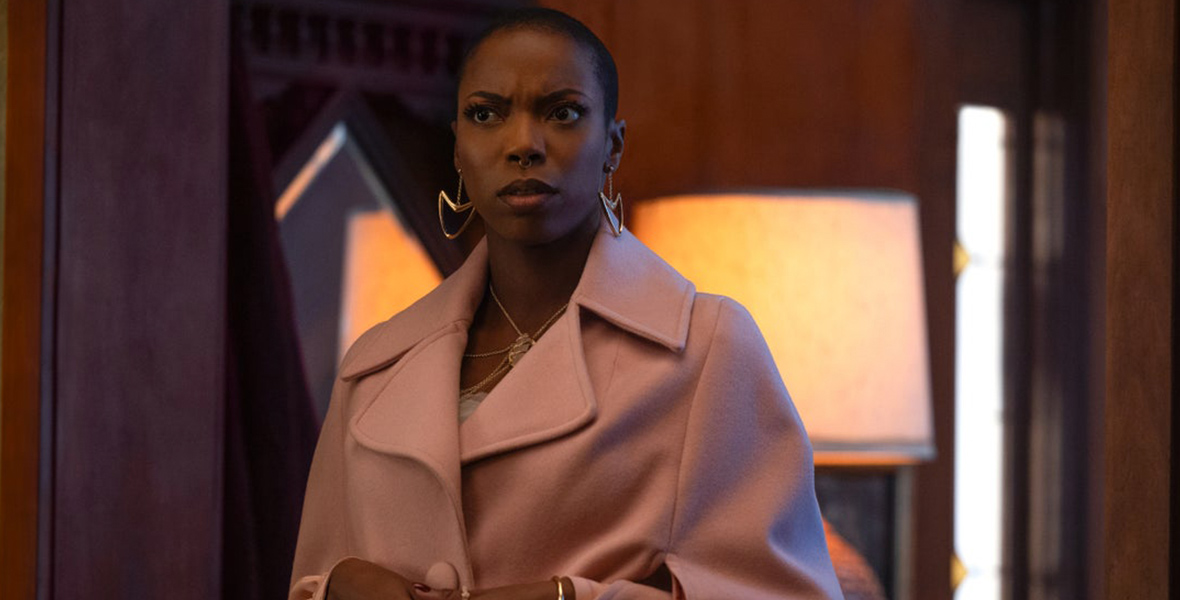 Jennifer Kale in a scene from Agatha All Along, Jennifer Kale (Sasheer Zamata) wears a rose-colored wool coat, along with gold earrings and a necklace. She has a confused expression on her face. The background includes a warmly lit coffee table lamp and a wooden interior.