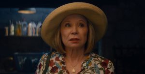 Mrs. Hart in a scene from Agatha All Along, Mrs. Hart (Debra Jo Rupp) is dressed in a floral flowy top, a gold necklace, and a straw hat. She looks to her left with raised eyebrows, wearing red lipstick. The background shows shelves stocked with laundry detergent and cleaning supplies.