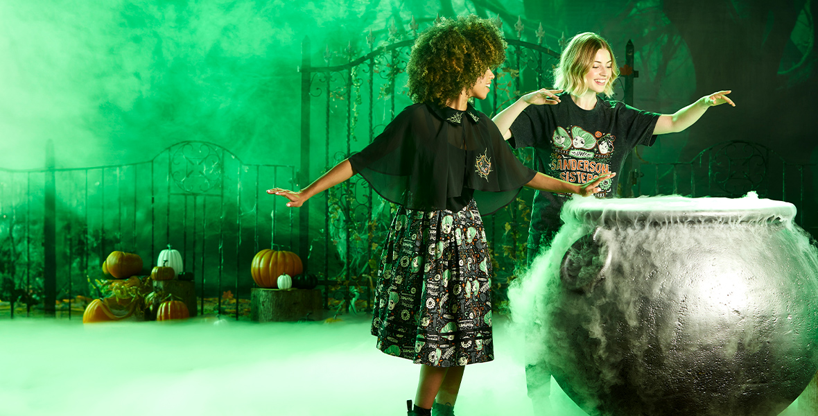 Two women are standing over an active cauldron leaking smoke, dressed in Hocus Pocus-themed clothing. One woman is wearing a two-piece dress with a chiffon capelet. The second woman is wearing a “Sanderson Sisters Live” t-shirt. They are smiling and have their arms raised over the cauldron as if they are conjuring a spell. In the background are black wrought iron gates, pumpkins, spooky trees, and foggy green hues.