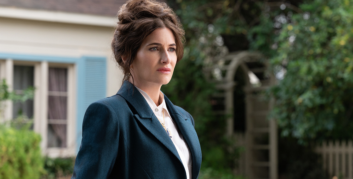 In a scene from Agatha All Along, Agatha Harkness (Kathryn Hahn) wears a navy blue fitted suit over a collared white shirt, and a gold pendant necklace. Her hair is in an updo, and the background features the exterior of a house and its garden.