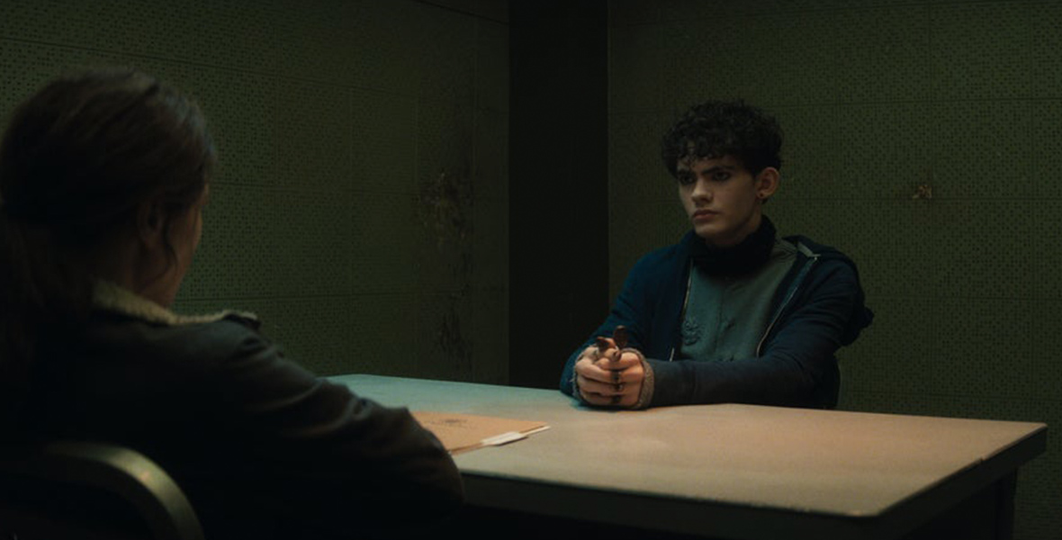 In a scene from Agatha All Along, Agatha Harkness (Kathryn Hahn) sits on a table opposite Teen (Joe Locke). His hands are laced on the table, Teen wears a gray sweater with a scrunched-up black mask around his neck, layered with a black zip-up cardigan. The background features the interior of a police station's interrogation room, with a file resting on the table.