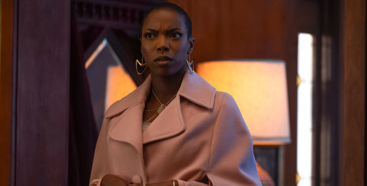 In a scene from Agatha All Along, Jennifer Kale (Sasheer Zamata) wears a rose-colored wool coat, along with gold earrings and a necklace. She has a confused expression on her face. The background includes a warmly lit coffee table lamp and a wooden interior.