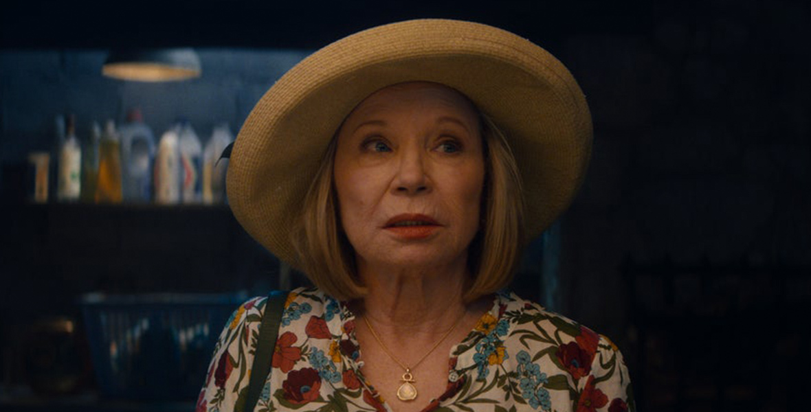 In a scene from Agatha All Along, Mrs. Hart (Debra Jo Rupp) wears a floral flowy top, a gold necklace, and a straw hat. She looks to her left with raised eyebrows, wearing red lipstick. The background shows shelves stocked with laundry detergent and cleaning supplies.