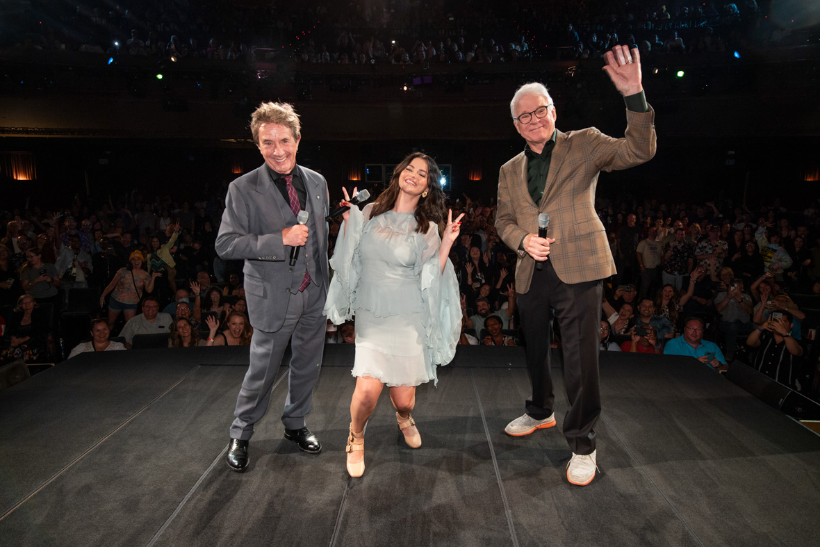 Martin Short, Selena Gomez, and Steve Martin onstage posing for a photo with the audience in the background. 