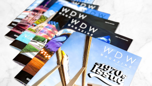 A collection of five WDW Magazine print issues fanned out across a marble table, with the top issue showing an image of the Walt Disney World Monorail and reading “The 1970s Issue.”