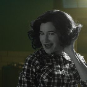 In a scene from Agatha All Along, Agatha Harkness (Kathryn Hahn) wears a retro, short hairstyle and a checkered shirt. She is smiling mischievously and posing with one hand in her hair.