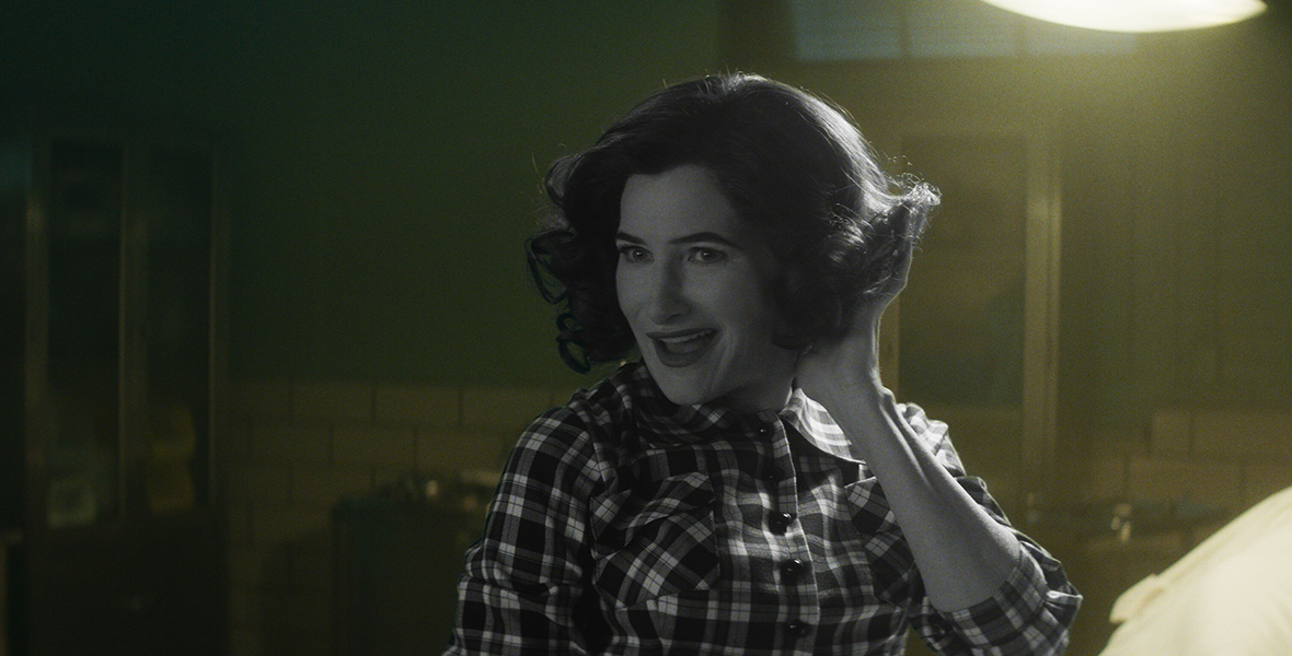 In a scene from Agatha All Along, Agatha Harkness (Kathryn Hahn) wears a retro, short hairstyle and a checkered shirt. She is smiling mischievously and posing with one hand in her hair.