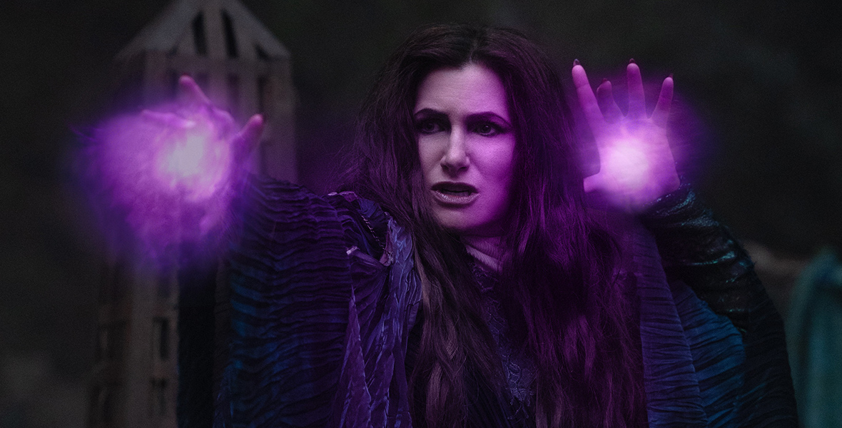 In a scene from Agatha All Along, Agatha Harkness (Kathryn Hahn) wears a long, dark purple velvet coat over a deep blue dress. Purple neon energy emanates from her hands as she gazes intensely ahead. The background features a tall gray tower against a dark, stormy sky.