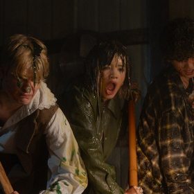 : In a first-look image from Carved (2024), (L-R) Corey Fogelmanis, Peyton Elizabeth Lee, and Wyatt Lindner, covered in colorful slime, hold shovels while digging. Their faces show shock and disgust. The dimly lit interior features wooden plank walls and metallic handles in the background.