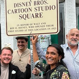 Jon Deutsch, Don Hahn, Justin Arthur, Braian Morrison, Nithya Raman, Roy P. Disney, Nick Runeare, and Joanna Miller posing with the Disney Bros. Cartoon Studio Square street sign. The sign reads: DISNEY BROS. CARTOON STUDIO SQUARE IN HONOR OF WALT AND ROY O. DISNEY, WHO OPENED THEIR FIRST STUDIO AT THIS LOCATION IN 1923.