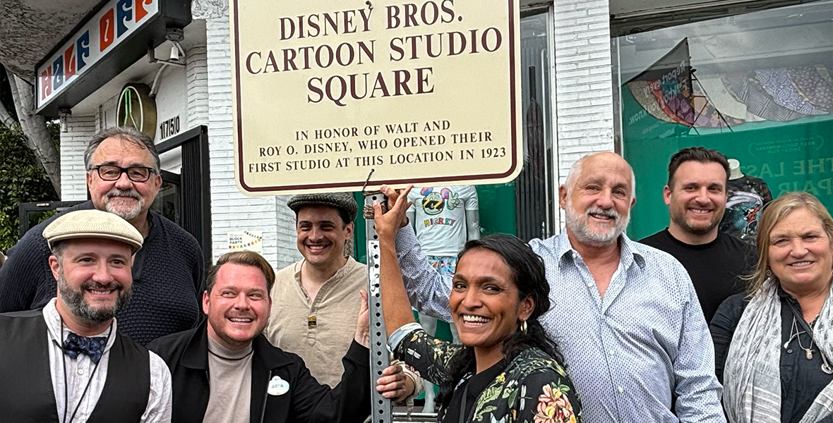 Jon Deutsch, Don Hahn, Justin Arthur, Braian Morrison, Nithya Raman, Roy P. Disney, Nick Runeare, and Joanna Miller posing with the Disney Bros. Cartoon Studio Square street sign. The sign reads: DISNEY BROS. CARTOON STUDIO SQUARE IN HONOR OF WALT AND ROY O. DISNEY, WHO OPENED THEIR FIRST STUDIO AT THIS LOCATION IN 1923.