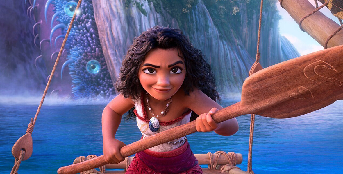 In an image from Walt Disney Animation Studios’ Moana 2, Moana is seen aboard her canoe in the middle of the ocean, holding an ore across her body with a determined look. In the background, there are greenery and waterfalls from a nearby island.